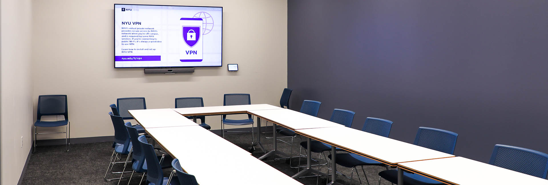 Conference room with rectangle table and blue chairs all around with white walls and a signature NYU purple accent wall and a flat screen TV in the front of the room on the wall of the NYU’s College of Arts and Sciences renovation by Structure Tone New York.