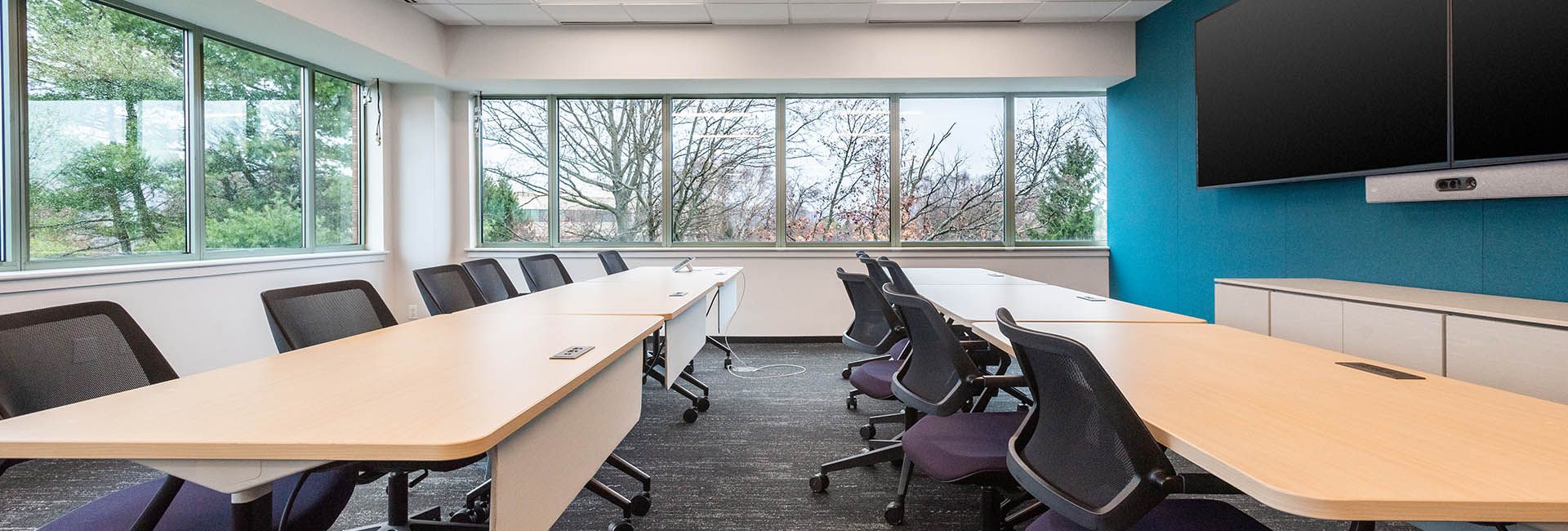 A conference room featuring a large screen and blue accent wall at the front and several chairs and desks arranged for attendees to view facing forward at an Accenture office.