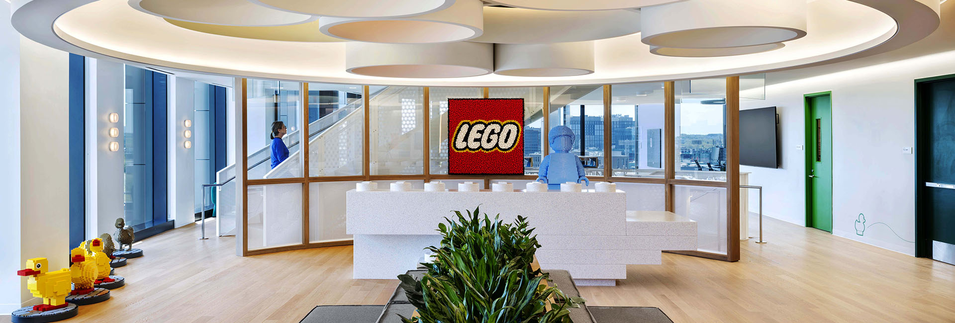 Reception area for LEGO Boston HQ built by Structure Tone Boston, colorful accent ceiling, benches with plants, Lego block reception desk and wall of windows.