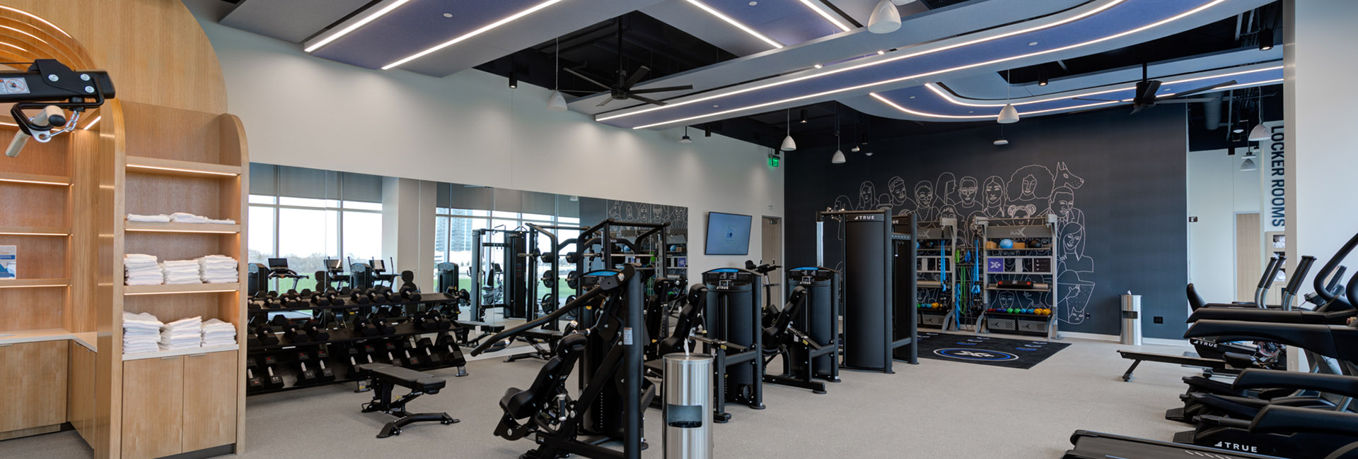 TIAA corporate center gym with intricate ceiling lighting blue accent wall weight equipment and treadmills along with wood towel rack built by Structure Tone Southwest