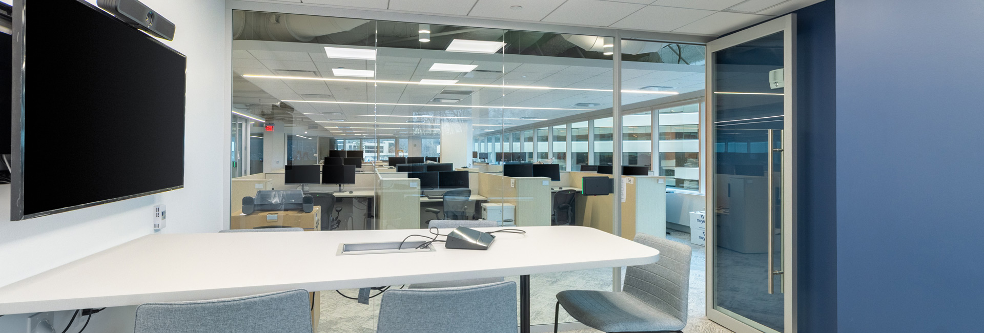 Inside a conference room with glass walls overlooking an open floor plan of cubicles