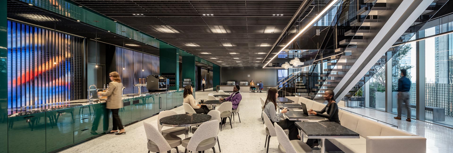 An office kitchen area featuring people seated at tables, engaged in conversation and enjoying their surroundings.