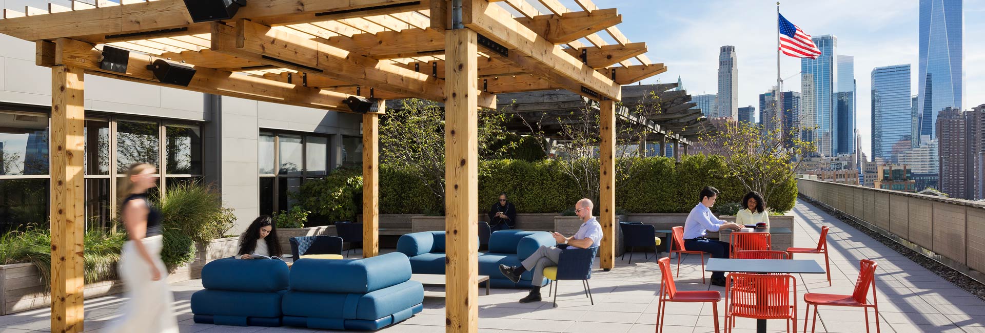 Rooftop terrace workspace with wooden pergolas, cushioned seating, and café tables, where employees sit and work outdoors with views of the city skyline and an American flag in the background.