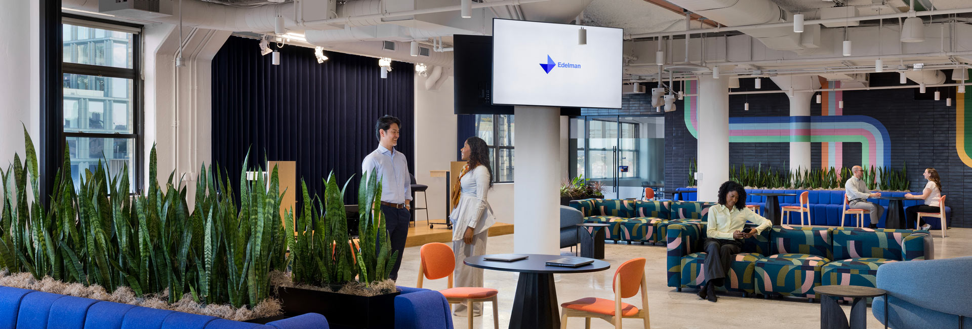 Modern office lounge with colorful seating, indoor plants, and employees talking, working, and relaxing beneath a large screen in an open, light-filled space.