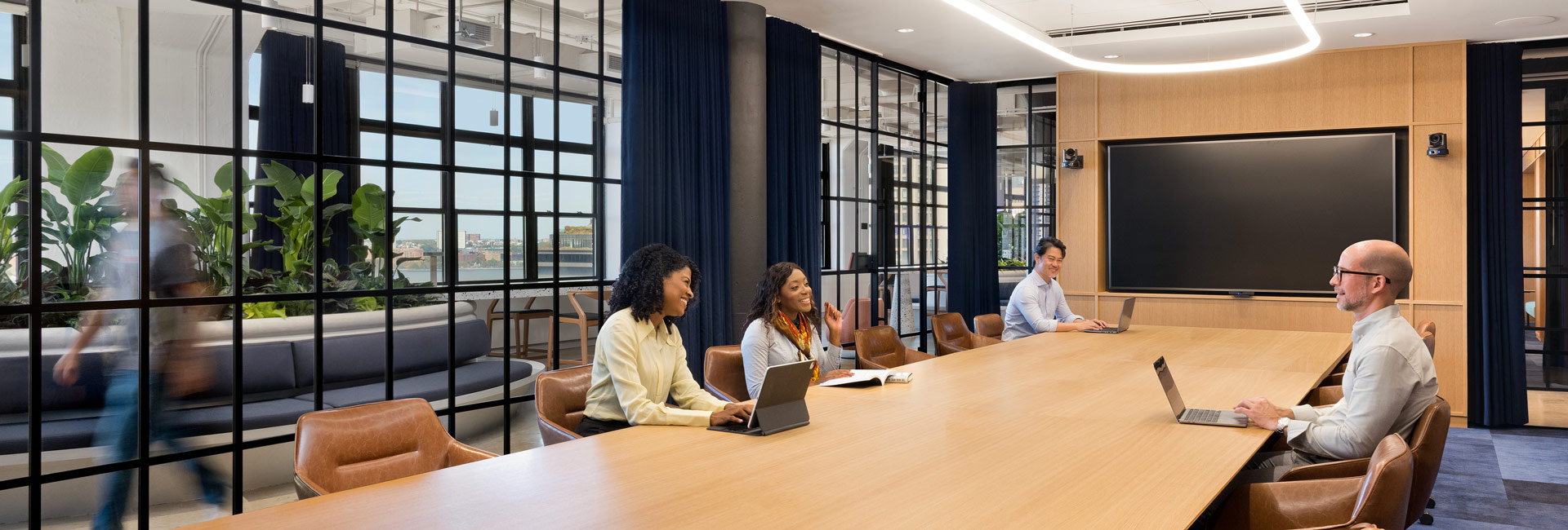 Modern conference room with a long wooden table, glass walls, and employees seated with laptops, collaborating in a bright, contemporary office setting.