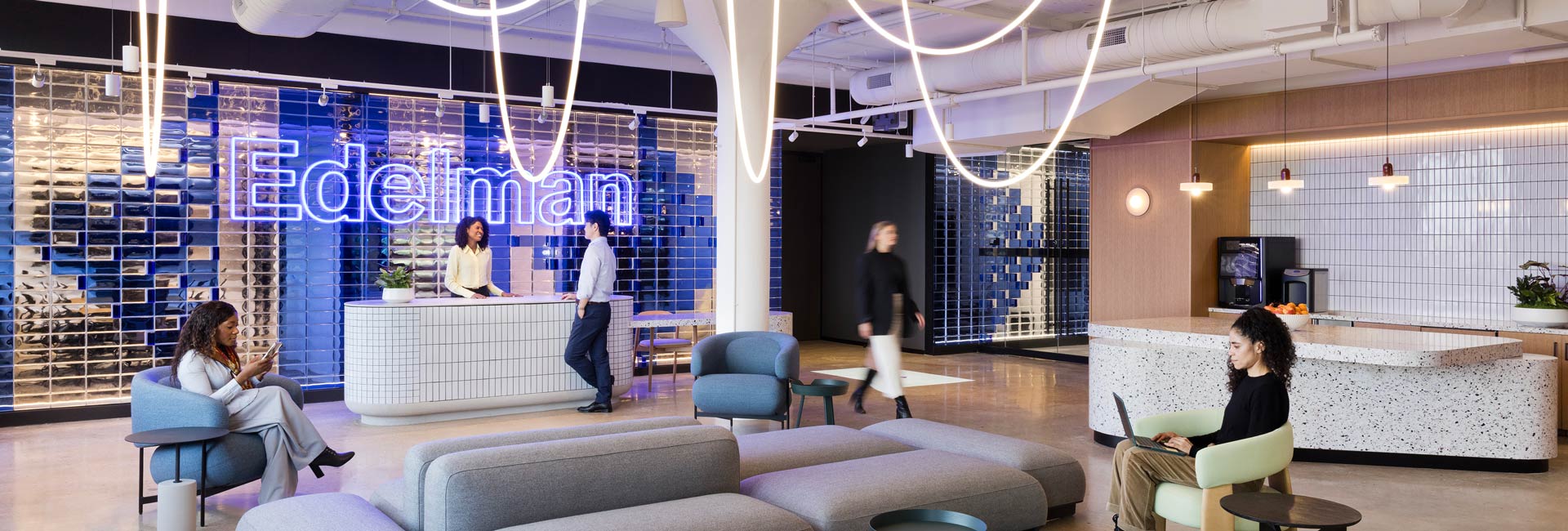 Modern office lobby with a reception desk, lounge seating, and a café-style counter, where employees are working, walking through the space, and checking their phones beneath sculptural lighting and a branded wall.
