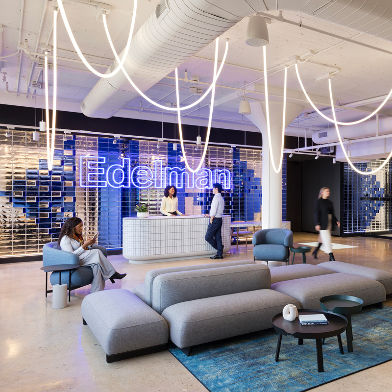 Modern office lobby with a reception desk, lounge seating, and a café-style counter, where employees are working, walking through the space, and checking their phones beneath sculptural lighting and a branded wall.