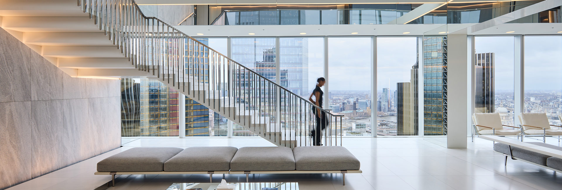A woman walking down a flight of stairs in an open office space overlooking the London skyline.