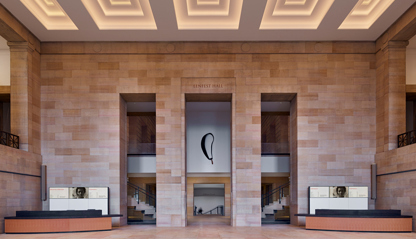 Grand stone-clad museum lobby with a coffered ceiling, symmetrical staircases, and a suspended black abstract sculpture centered between three tall openings.