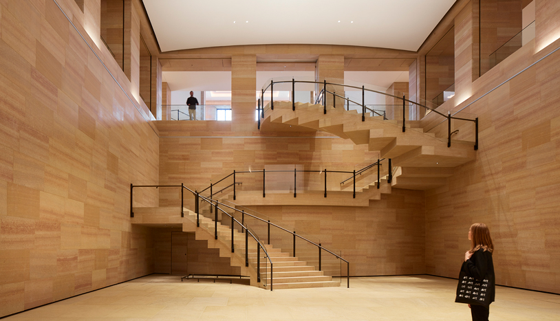 Large, light-filled interior atrium clad in warm beige stone, featuring a dramatic multi-level staircase with zigzagging flights and glass-and-metal railings.