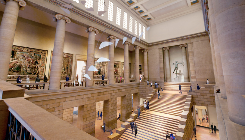 Grand museum hall with soaring stone columns, high clerestory windows, and a broad central staircase leading between gallery levels.