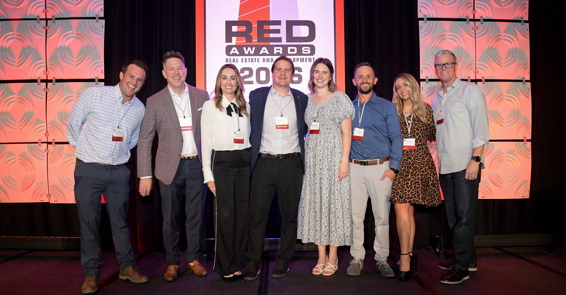 Eight conference attendees pose together on a stage in front of a large RED Awards backdrop during a real estate and development industry event.