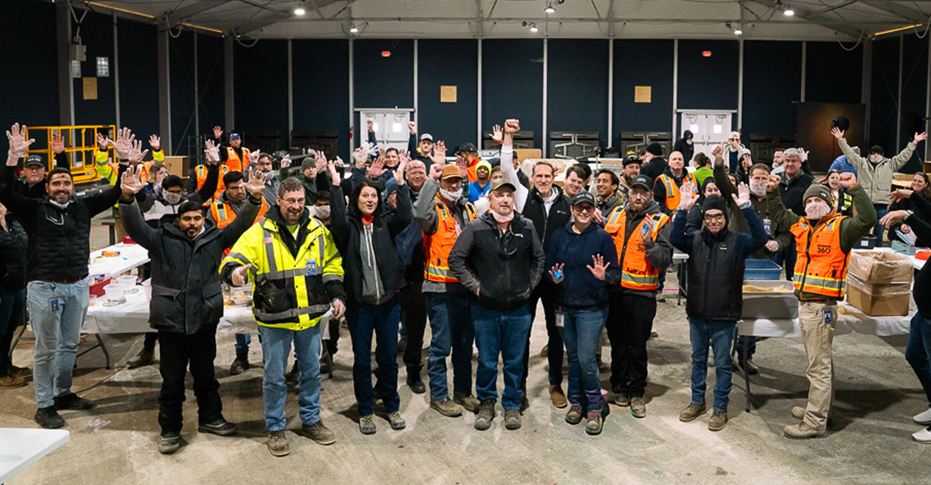 A large group of about 40–50 people stand inside a warehouse or industrial facility, facing the camera and waving.
