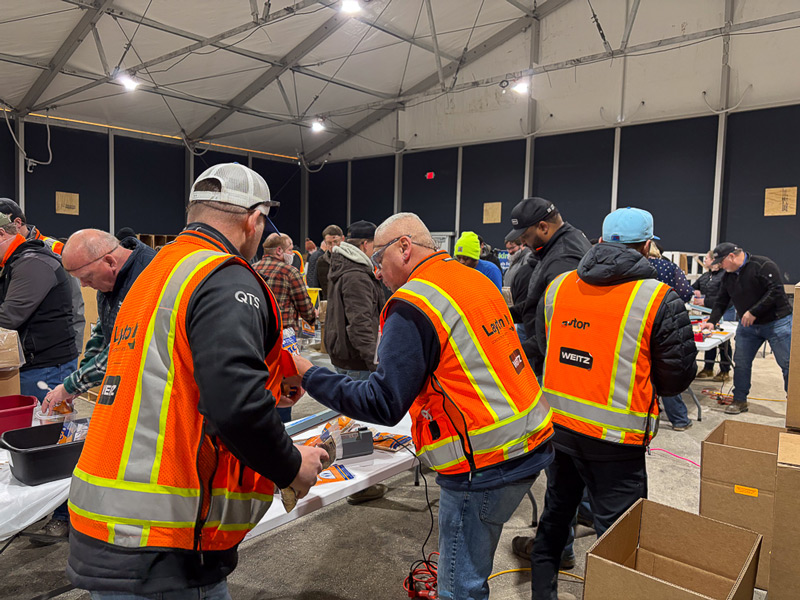 A group of construction and project workers wearing orange high-visibility safety vests gather around tables inside a large temporary tent structure. They are assembling or sorting items for a volunteer event, with boxes, tools, and supplies spread across the tables.
