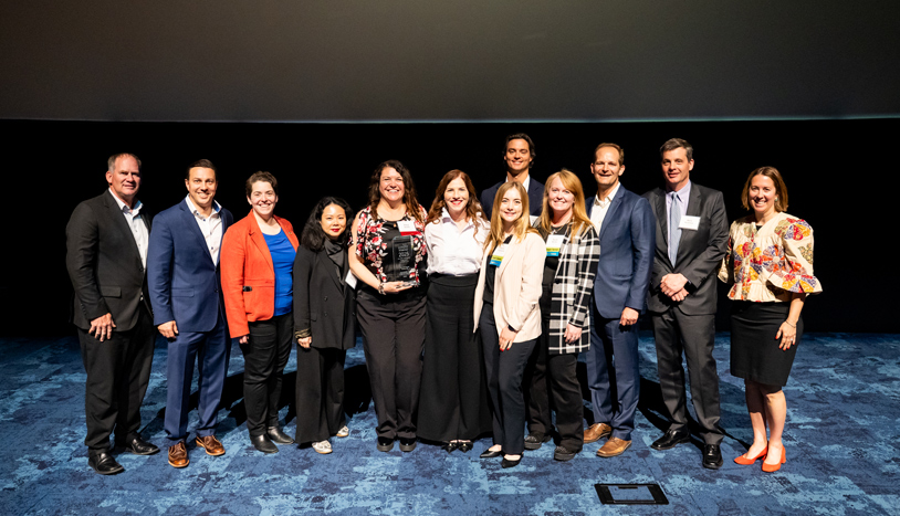 A group of professionals poses on stage as a team member holds an award trophy, celebrating their recognition for winning an industry award.
