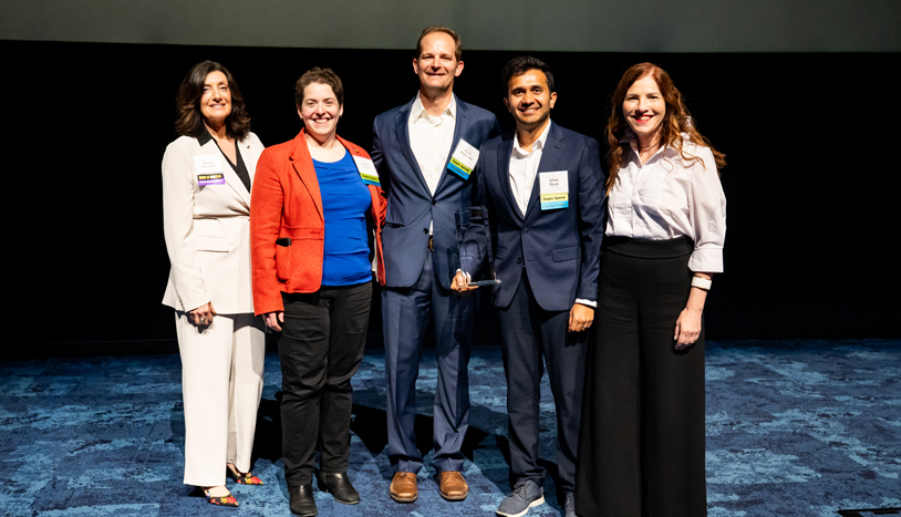 A group of professionals poses on stage as a team member holds an award trophy, celebrating their recognition for winning an industry award.