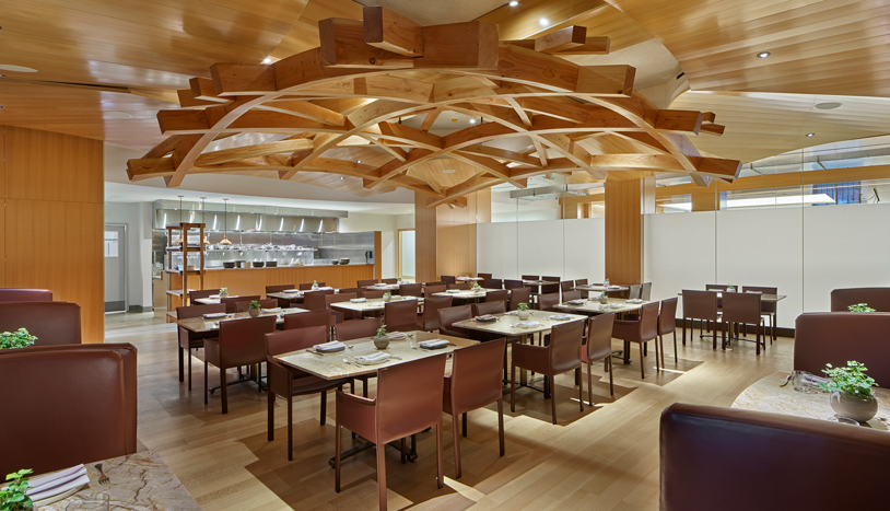 Warmly lit restaurant dining room with wood-paneled walls and ceiling, neatly arranged tables and chairs, and a sculptural wooden lattice canopy suspended above the seating area.