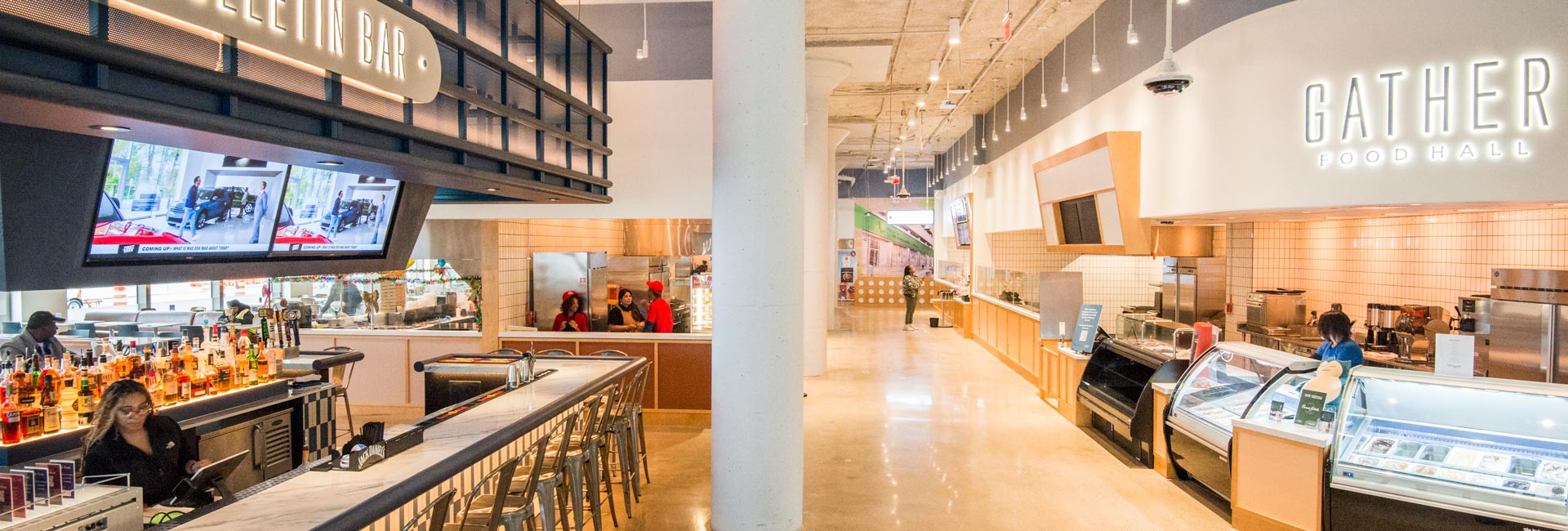 Interior view of a modern food hall featuring a central bar with televisions, open food vendor counters, bright lighting, and wide walkways.