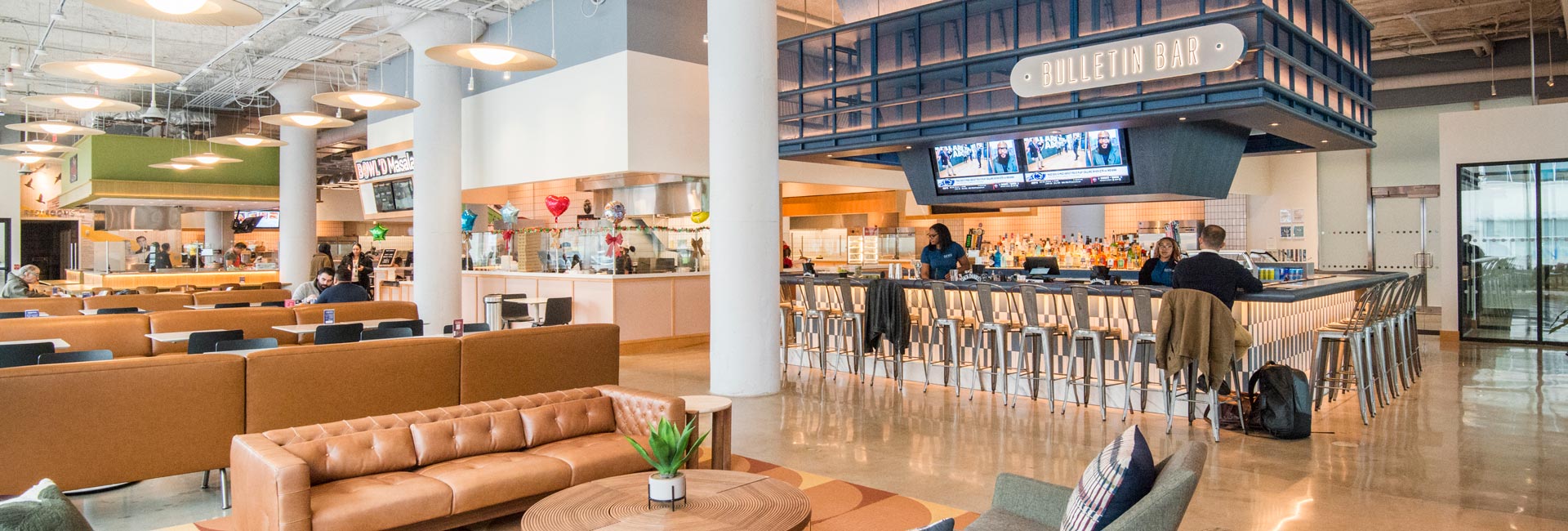 Spacious food hall with lounge seating, communal dining tables, a large bar area, and multiple food vendors in an open-concept setting.