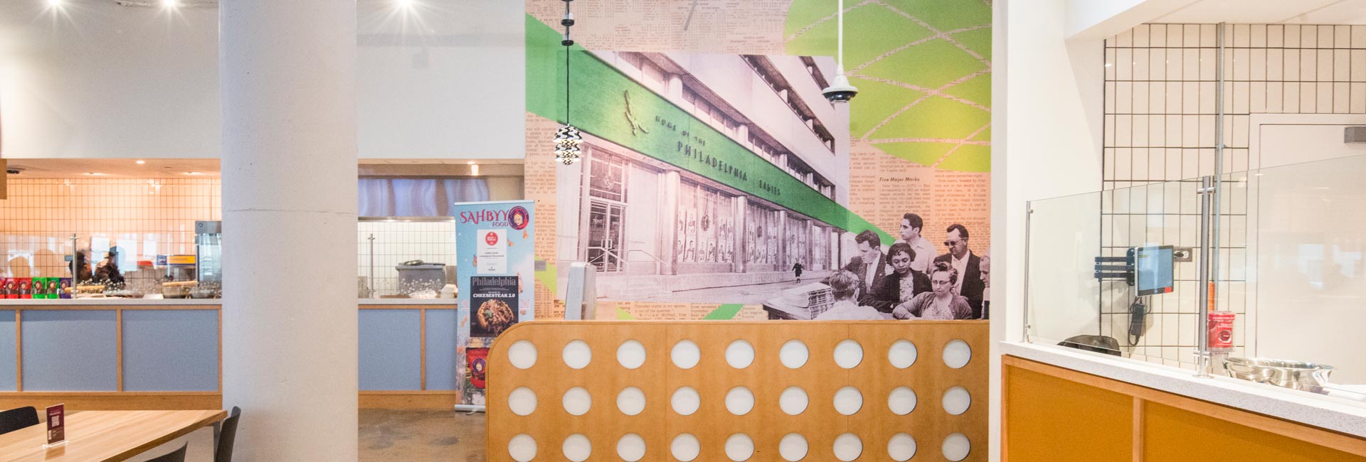 Dining space within a food hall showcasing a large historical mural, wooden tables, food vendor counters, and a bright, open atmosphere.