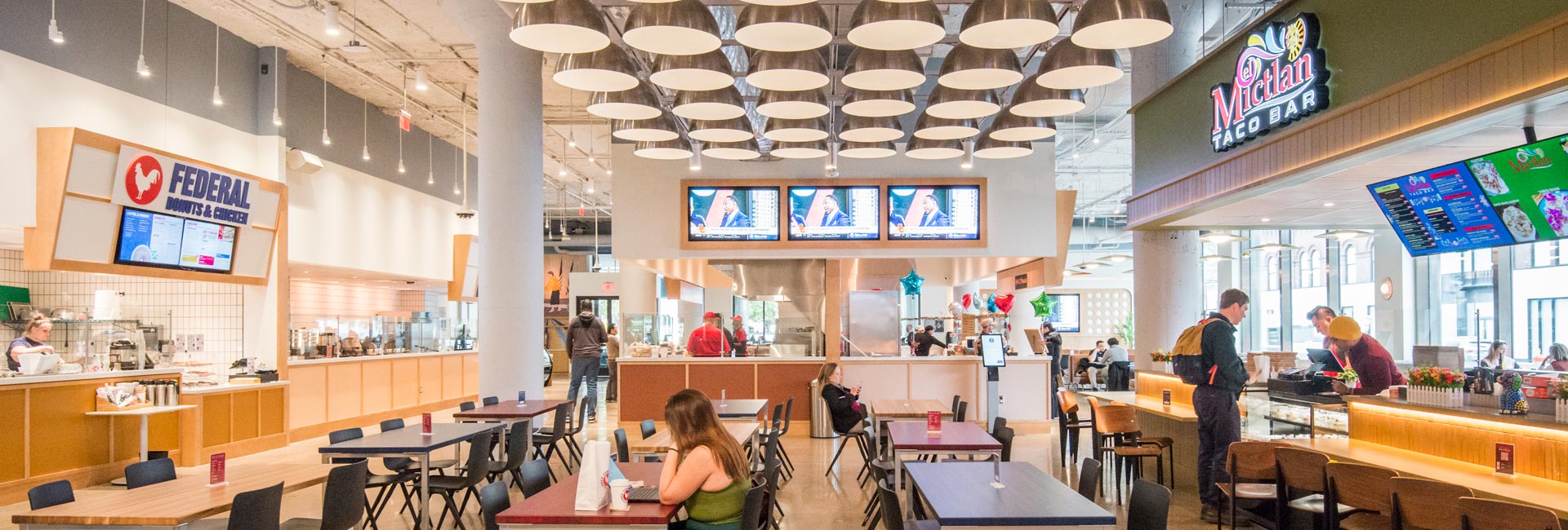 Interior view of a modern food hall featuring a central bar with televisions, open food vendor counters, bright lighting, and wide walkways.
