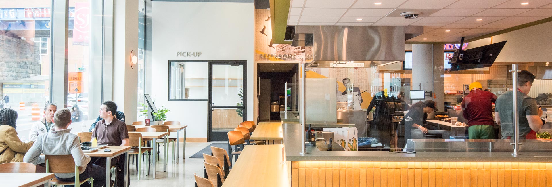 Interior view of a modern food hall featuring a central bar with televisions, open food vendor counters, bright lighting, and wide walkways.