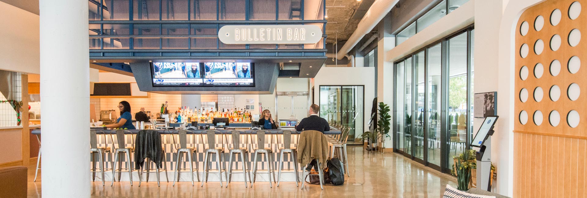 Interior view of a modern food hall featuring a central bar with televisions, open food vendor counters, bright lighting, and wide walkways.