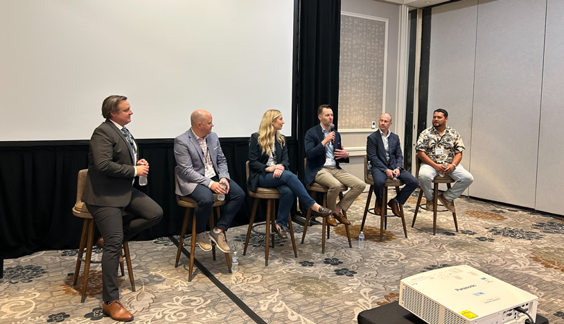 Six panelists seated on tall stools participate in a discussion in a conference room, with one speaker holding a microphone while addressing the audience.
