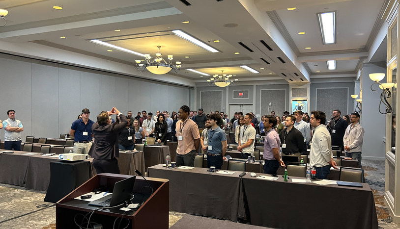Participants stand among conference tables in a hotel meeting room while an instructor leads an interactive group activity at the front of the session.