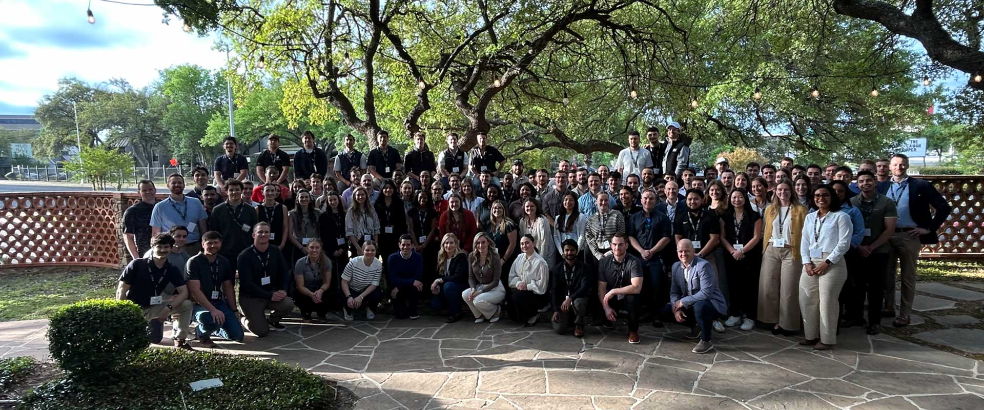 Large group of people posing for a team photo on an outdoor stone patio beneath sprawling trees strung with lights, with greenery and a low decorative wall in the background.