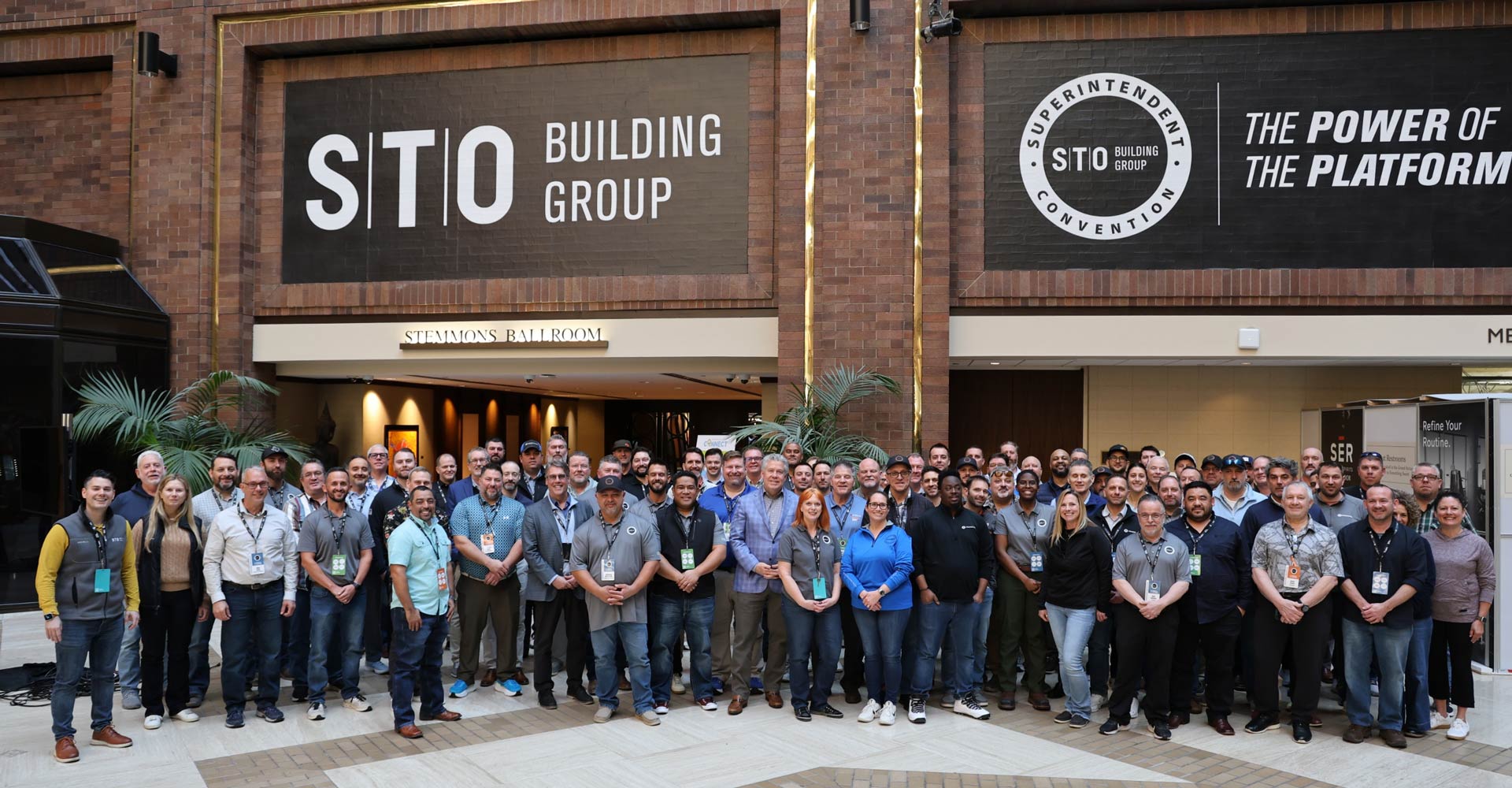 Large group of conference attendees posing for a photo in a hotel atrium beneath banners for STO Building Group and the Superintendent Convention.