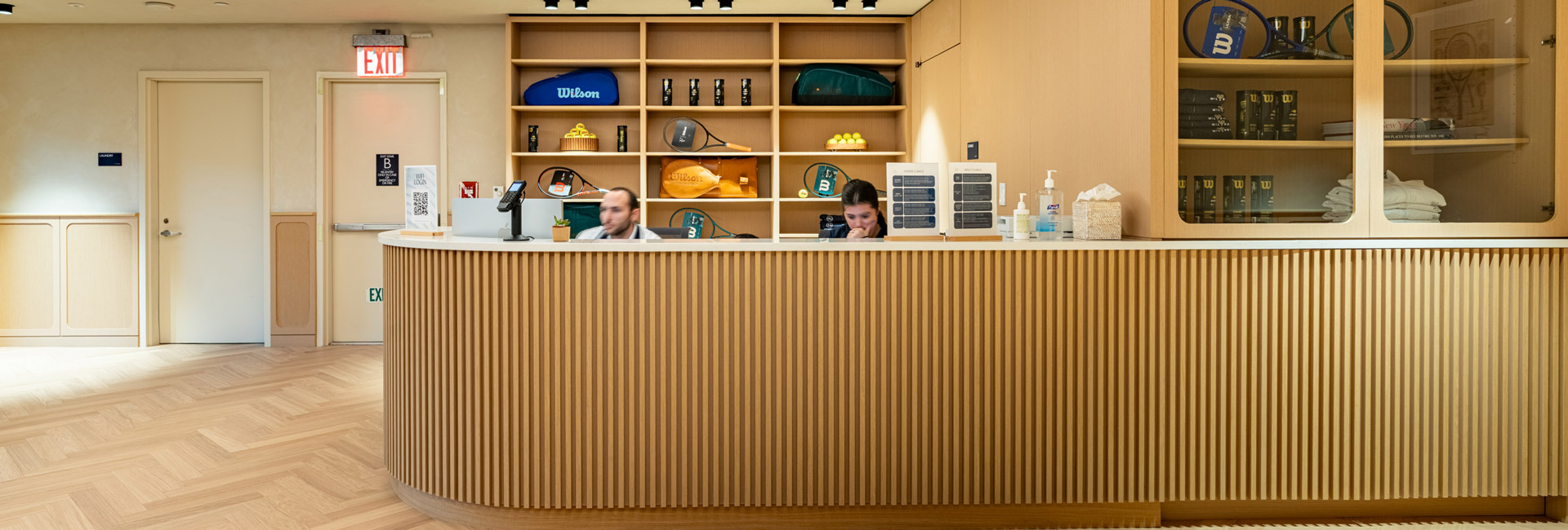 Wood-paneled reception desk with staff and displays of tennis racquets, balls, and accessories.