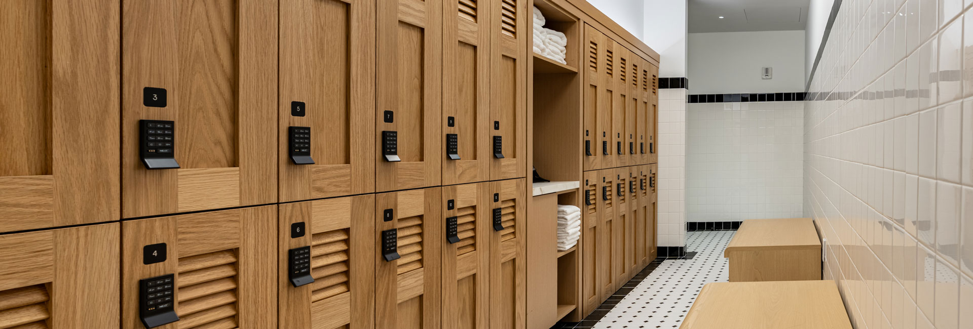 Locker room with numbered wood lockers, integrated benches, towel storage, and white tile walls.