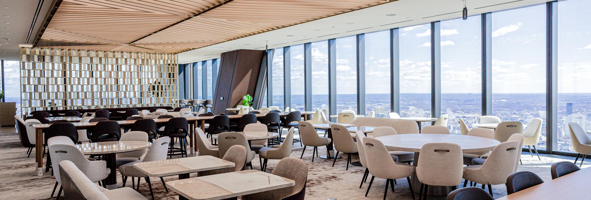 Bright corporate dining and gathering space with varied seating, a slatted wood ceiling, and floor-to-ceiling windows overlooking the city.