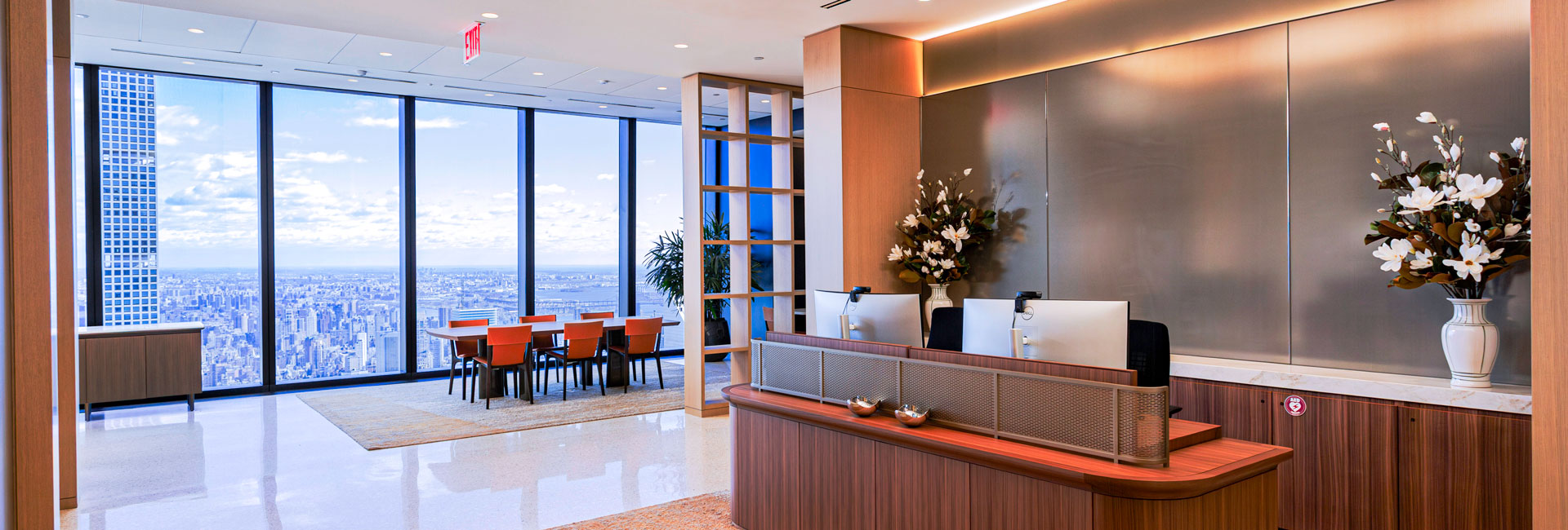 Executive reception area with a curved wood desk, floral arrangements, adjacent meeting space, and expansive city views.