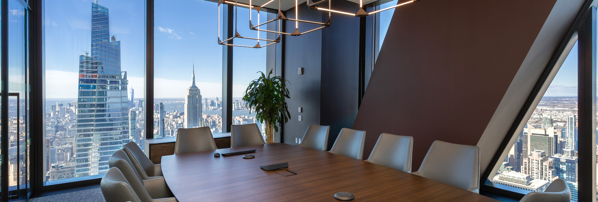 Modern conference room with a wood table, white chairs, geometric lighting, and panoramic views of the Manhattan skyline.