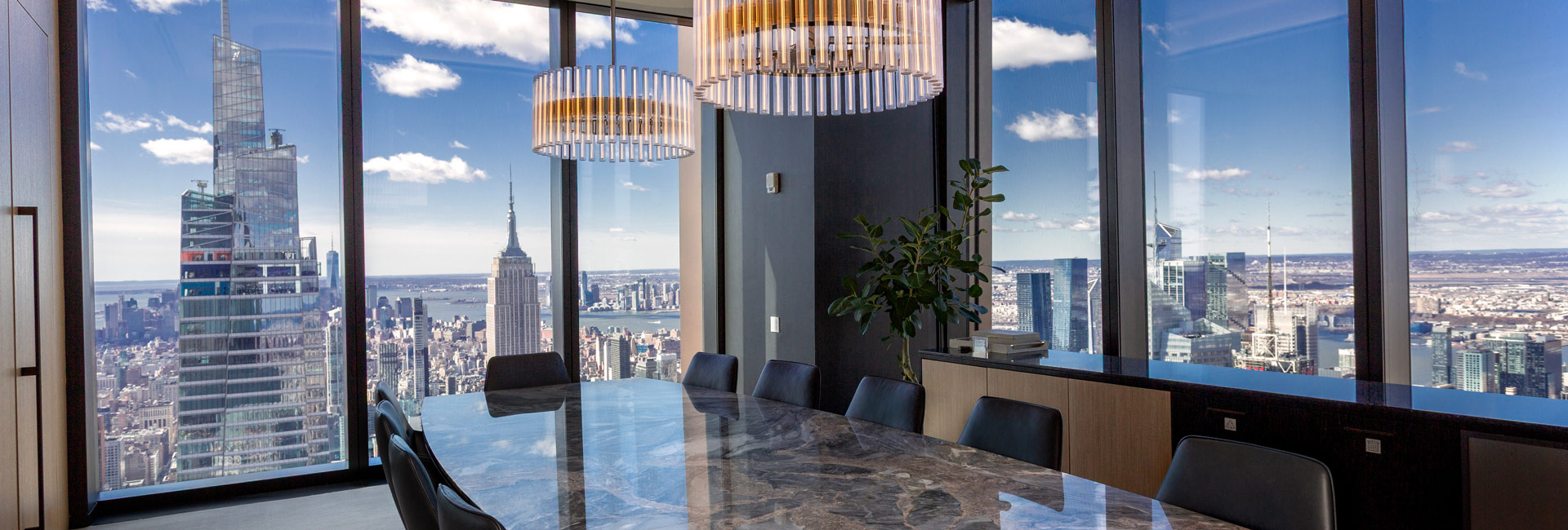 Boardroom with a polished stone table, black chairs, statement chandeliers, and expansive views of the Empire State Building and Manhattan skyline.