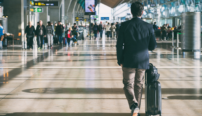 Provide alt text for this image. Business traveler pulling a suitcase through a busy, sunlit airport terminal.