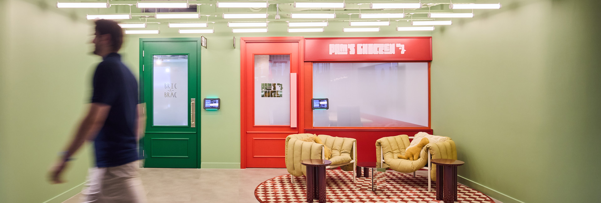 Colourful office meeting rooms styled as high-street shops, including a red chicken shop frontage and green door, with lounge seating outside.