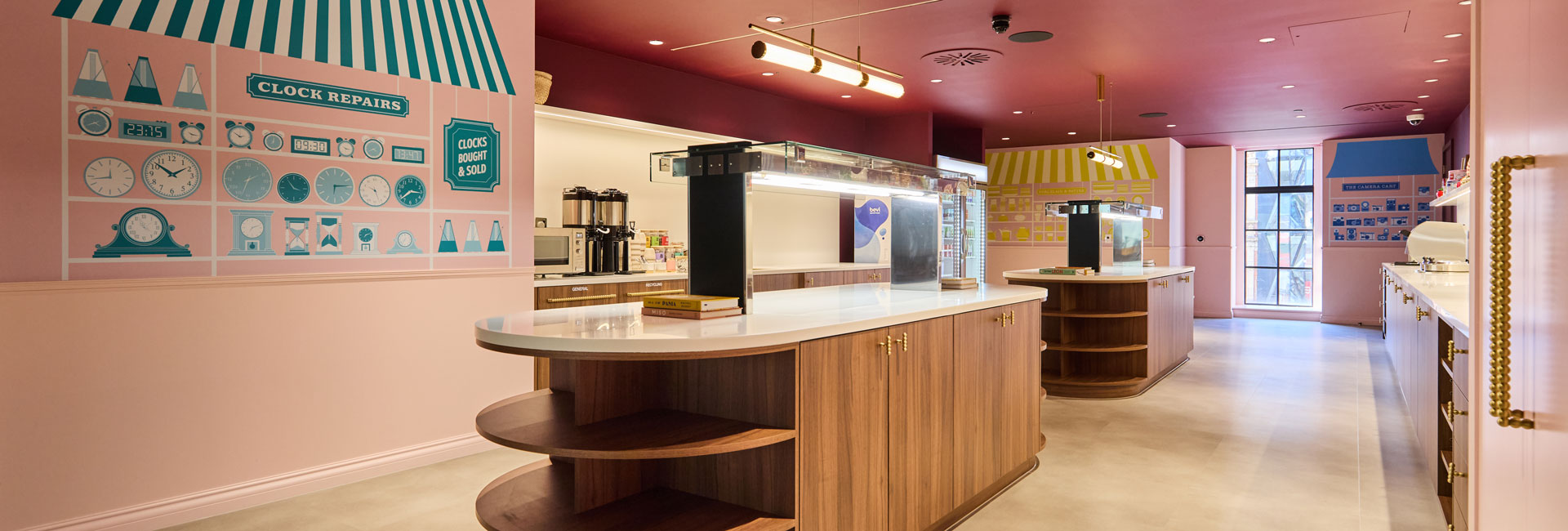 Pink office dining area with curved wooden counters, brass details and illustrated clock repair and camera shop murals.