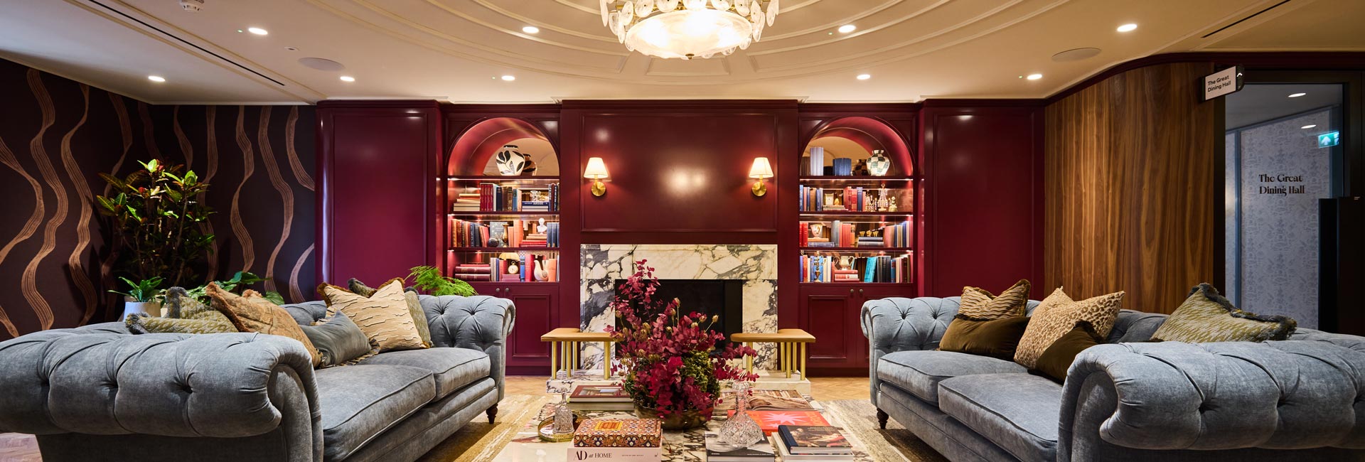 Drawing room with burgundy panelling, marble fireplace, bookcases and two grey tufted sofas beneath a chandelier.