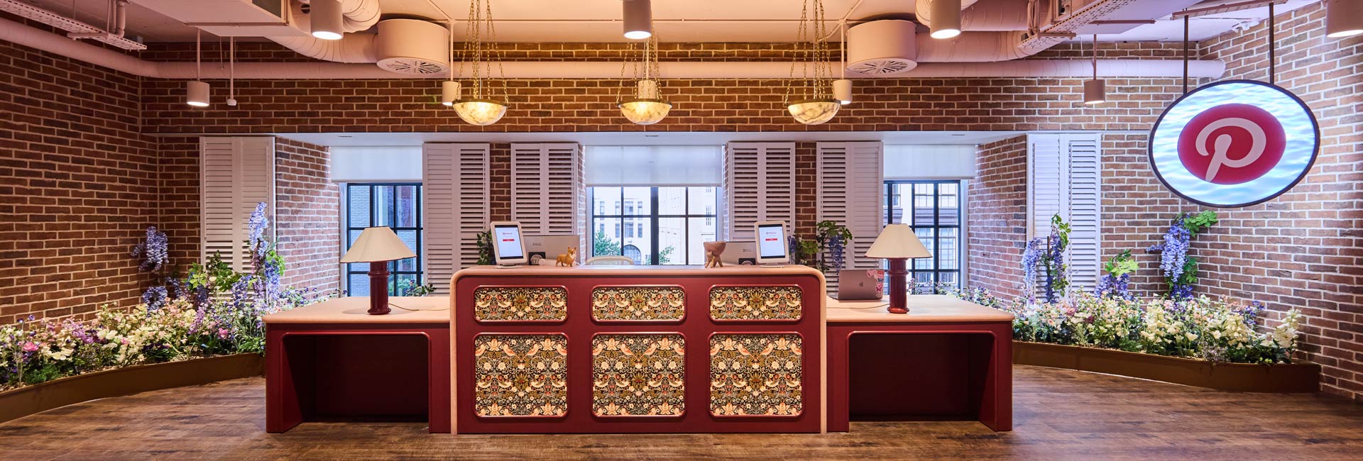 Pinterest reception desk framed by exposed brick walls, floral planting, shuttered windows and an illuminated Pinterest logo.