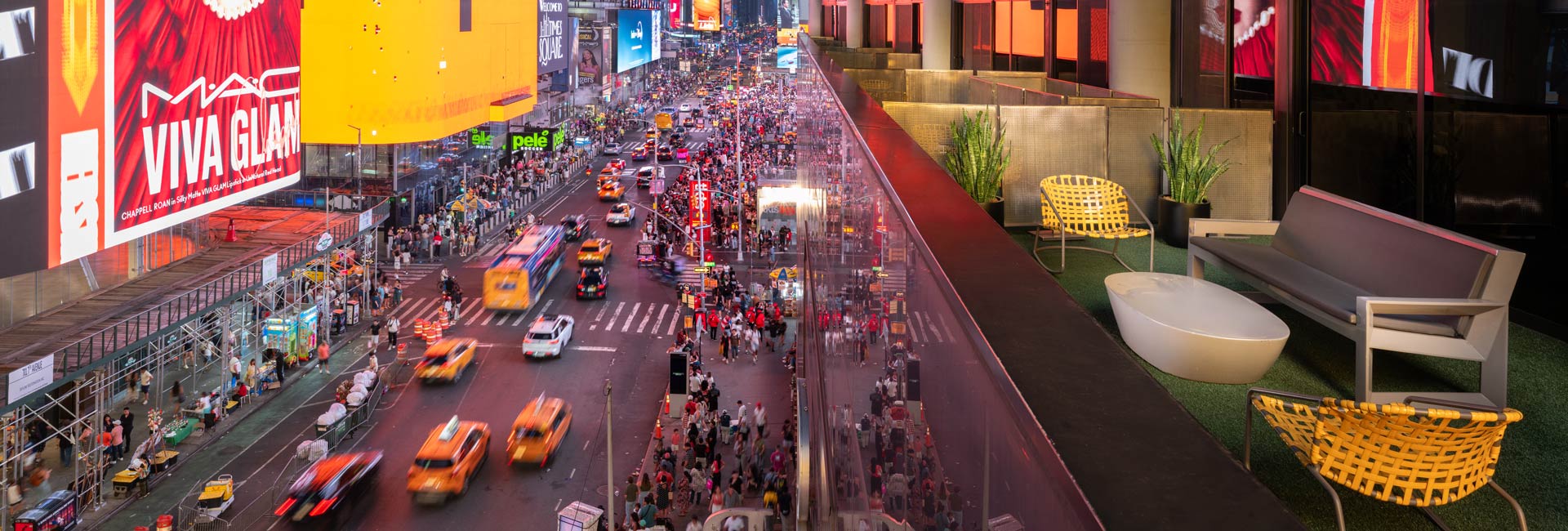 Furnished hotel balcony overlooking the bright billboards, traffic, and crowds of Times Square at dusk.