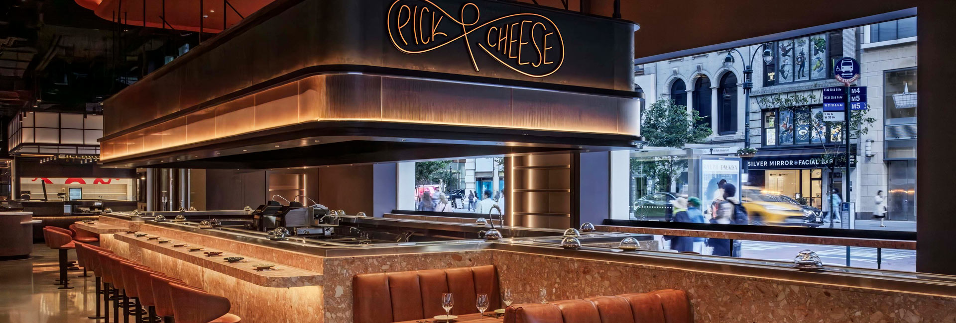 Pick & Cheese counter with marble surfaces, leather seating, warm lighting, and street views through large windows.