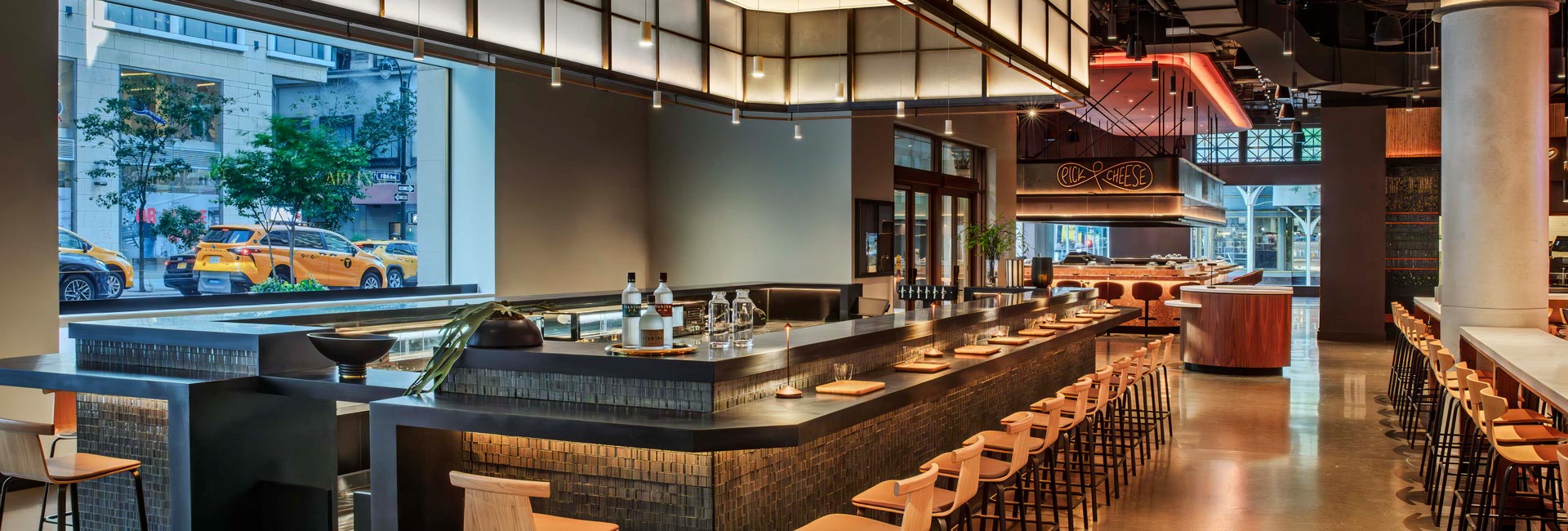Modern bar with tiled surfaces, wood stools, pendant lighting, and floor-to-ceiling windows overlooking the street.