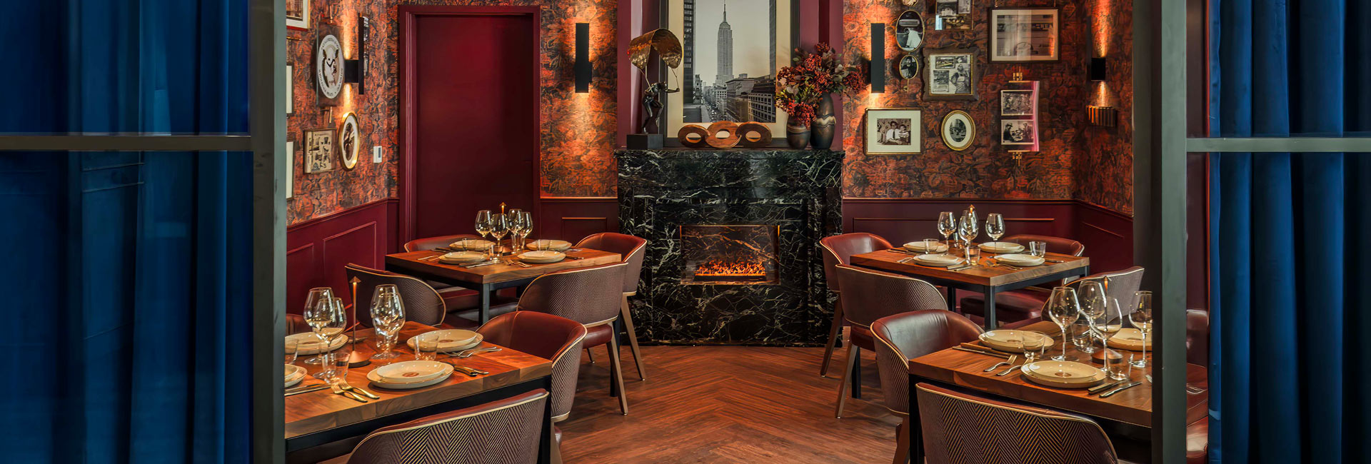 Intimate dining room with set tables, rich red walls, framed artwork, and a black marble fireplace.