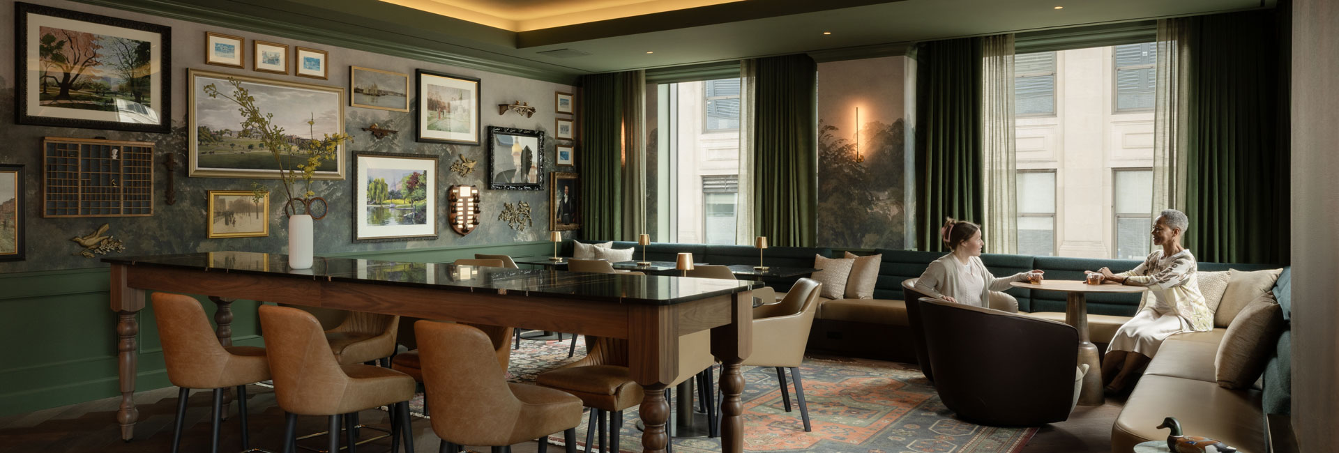 Library-inspired workplace lounge with gallery walls, green banquette seating, communal tables, and two colleagues talking.