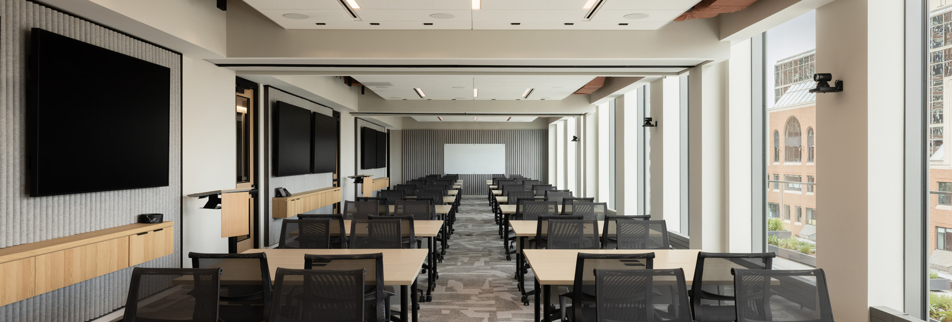 Sunlit training room with modular tables, rows of chairs, presentation screens, and floor-to-ceiling windows.