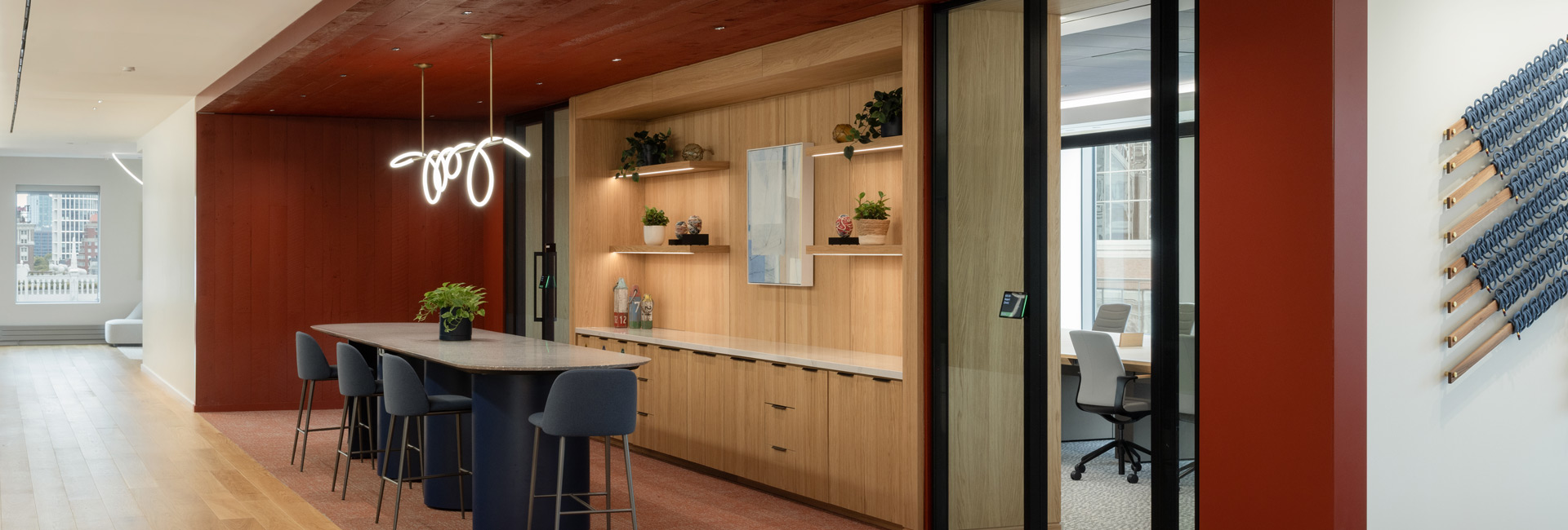 Office collaboration area with a high table, blue stools, warm wood cabinetry, red accents, and an adjacent glass meeting room.