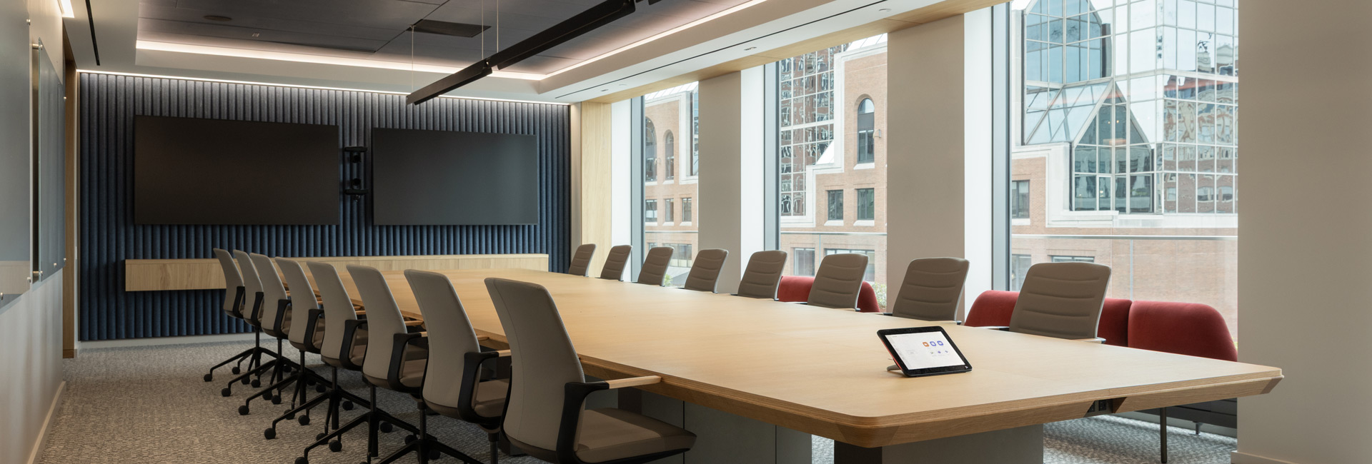 Modern boardroom with a large wood conference table, dual presentation screens, and floor-to-ceiling windows overlooking Boston.